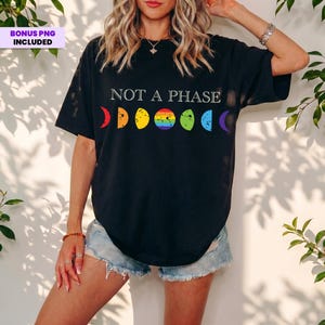 May include: A black t-shirt featuring the white text NOT A PHASE above a horizontal row of moon phases. Each moon phase is colored in a different shade of the rainbow, including red, orange, yellow, green, blue, and purple, with a full rainbow circle.