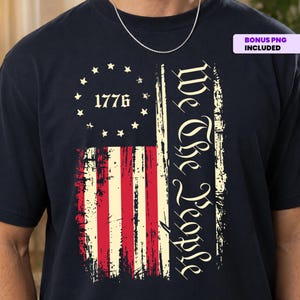 Puede incluir: Una camiseta azul marino oscuro con un gráfico desgastado de la bandera estadounidense, trece estrellas en círculo, el año 1776 y el texto We The People escrito en una fuente vertical estilizada. El diseño tiene una apariencia envejecida.
