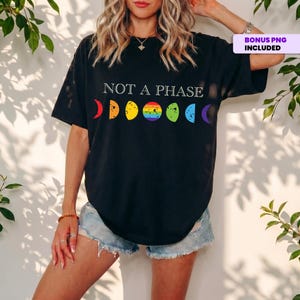 Op de afbeelding: Zwart T-shirt met de tekst "NOT A PHASE" boven een regenboogkleurige afbeelding van de maanfasen. Het shirt is donker van kleur en heeft een relaxte pasvorm. Het ontwerp is een statement van steun.