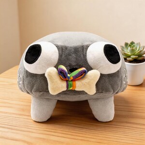 May include: A gray plush toy shaped like a rounded creature with two large white eyes featuring black pupils. The creature holds a cream-colored bone-shaped toy in its mouth, which is tied with a small, vibrant, multi-colored rainbow ribbon.