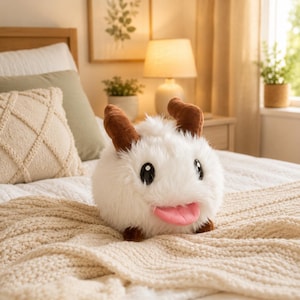 May include: A plush white creature with brown horns, black eyes, and a pink tongue. The stuffed animal is on a cream-colored blanket. The plush toy is a cute and cuddly character.