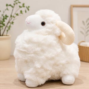 May include: A plush sheep toy with soft white faux fur, a smooth white face, dark brown eyes, and a small pink stitched mouth. The stuffed animal features two curved, ribbed, cream-colored horns on the sides of its head and four white paws.