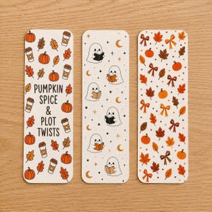 May include: Three rectangular bookmarks with rounded corners. The left one features pumpkins, coffee cups, and the text PUMPKIN SPICE & PLOT TWISTS. The center one shows ghosts reading books with stars and moons. The right one displays autumn leaves and bows.