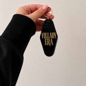 May include: A black motel-style keychain with a diamond shape and rounded edges. The keychain features the text "VILLAIN ERA" printed in gold lettering and includes a gold-colored metal ring attached to the top for holding keys.