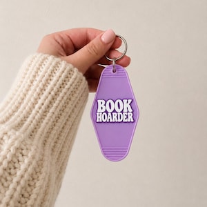 May include: A lavender-colored motel-style keychain with a diamond-shaped body and horizontal ridges at the top and bottom. The text BOOK HOARDER is embossed in white, bold, sans-serif capital letters in the center of the keychain.