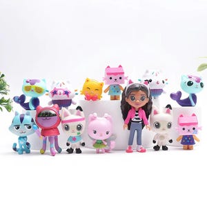 May include: A collection of colorful Gabby's Dollhouse figurines, including cat-themed characters with various outfits and accessories. The figures are in different colors, such as blue, pink, yellow, and white, and are arranged on a white surface.