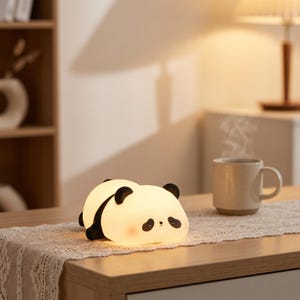 以下が含まれることがあります： 黒い耳とアクセント、かわいらしい眠そうな表情が特徴の、柔らかく光るパンダ型のナイトライト。木製のテーブルの上にあり、近くにはコーヒーカップがあります。どんな部屋にも魅力的なパンダライトです。