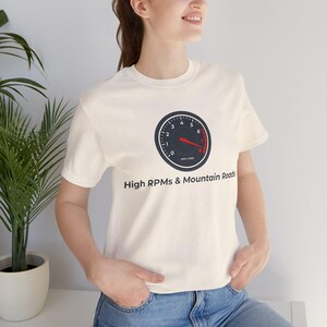 May include: A cream-coloured short-sleeve t-shirt featuring a graphic of a black tachometer gauge with a red needle pointing towards the high RPM range. Below the gauge, the text reads High RPMs & Mountain Roads in a simple black font.