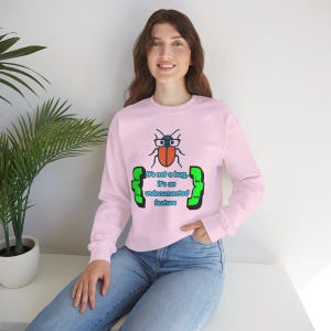 May include: A light pink long-sleeve sweatshirt featuring a graphic of a beetle wearing glasses. Below the beetle, the text reads, "It's not a bug, it's an undocumented feature," framed by two bright green curly brackets on either side.