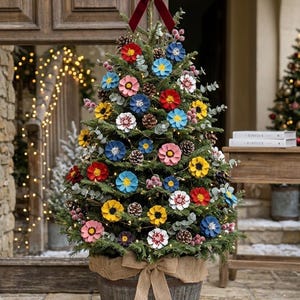 May include: A tabletop evergreen tree in a wooden barrel wrapped with a burlap bow. The tree is decorated with painted pinecone flowers in red, blue, yellow, pink, and white, along with natural pinecones, berries, and a dark red velvet bow topper.