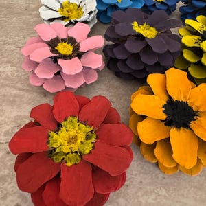 May include: A collection of pinecones painted to resemble flowers in various colors including red, orange, yellow, pink, purple, blue, and white. Each pinecone features painted petals and a textured center, creating a floral appearance.