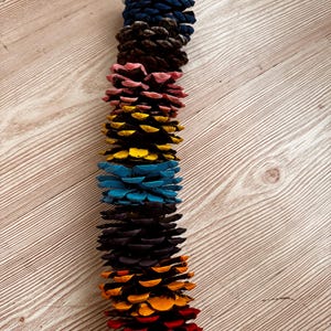 May include: A vertical garland composed of multiple pine cones, each painted in a distinct solid color. The sequence features silver, dark blue, brown, pink, yellow, light blue, dark purple, orange, and red pine cones strung together in a line.