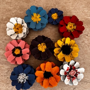 May include: A collection of ten pinecone flowers painted in various colors including blue, white, red, yellow, pink, dark purple, orange, and navy. Each pinecone is arranged to resemble a flower with a distinct center, showcasing vibrant hues.