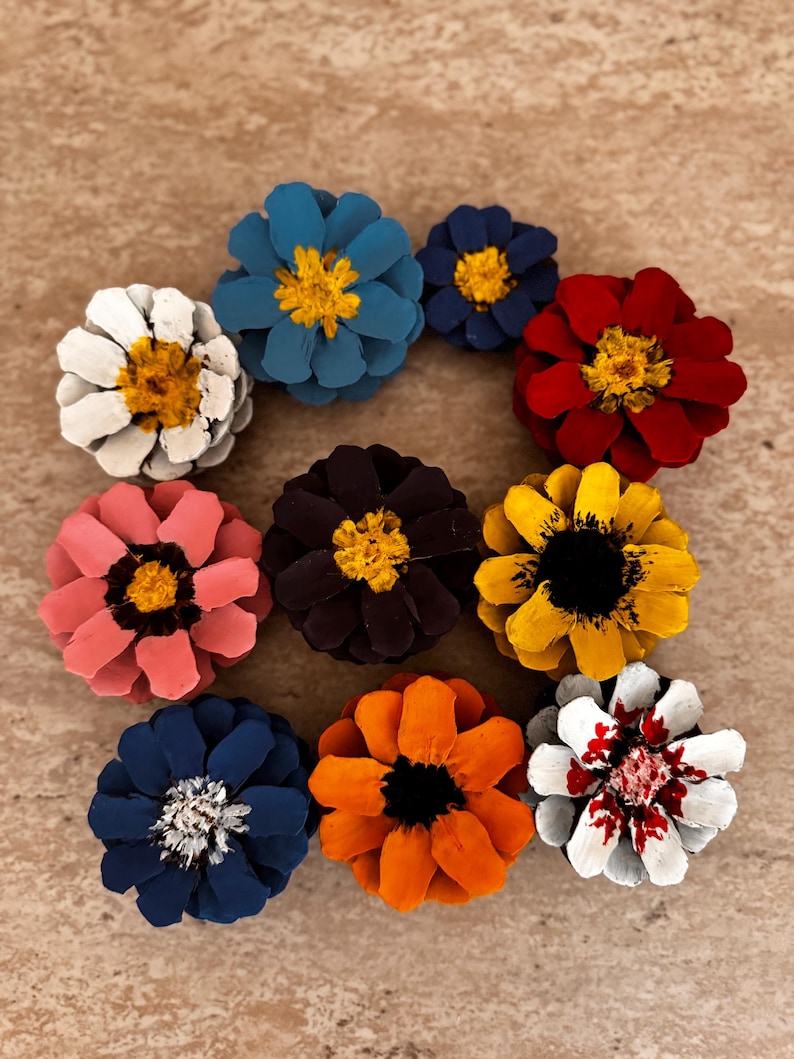 May include: A collection of ten pinecone flowers painted in various colors including blue, white, red, yellow, pink, dark purple, orange, and navy. Each pinecone is arranged to resemble a flower with a distinct center, showcasing vibrant hues.