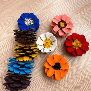 May include: A collection of pinecones painted to resemble flowers. The arrangement includes individual pinecones painted in solid blue, pink, red, orange, white, yellow, and purple, with some featuring contrasting centers in black, yellow, or white.