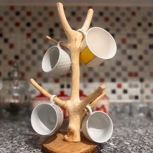 May include: A light-colored wooden mug tree with a circular base and multiple branches for hanging cups. Four ceramic mugs are suspended on the branches, including one with a yellow band and others with white textured or patterned surfaces.