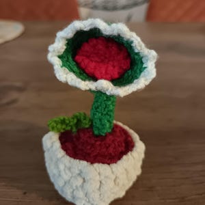 Peut inclure: Une figurine de plante crochetée présentant une tête de fleur à bouche ouverte rouge, verte et blanche sur une tige verte. La plante est placée dans un pot en crochet blanc texturé rempli de laine rouge foncé, avec une petite feuille verte.