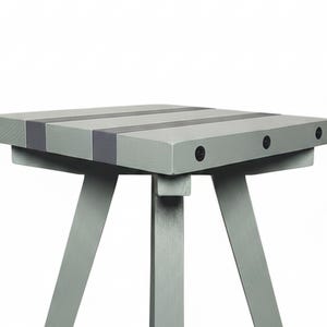 May include: A light gray wooden stool with a square seat featuring darker gray stripes. The stool has four angled legs connected by horizontal support braces and is finished with black hardware accents at the joints and black feet at the base.