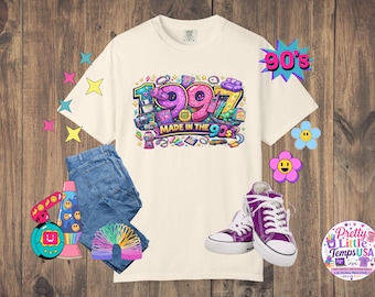 Könnte beinhalten: Ein cremefarbenes T-Shirt mit einer bunten Grafik mit der Aufschrift 1997 MADE IN THE 90S, umgeben von Abbildungen von Retro-Gegenständen wie einem Kaugummiautomaten, einem Pager und einem Handheld-Spiel, kombiniert mit blauer Jeans und lila Turnschuhen.