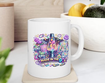 May include: A white ceramic mug featuring a colorful graphic illustration of 1990s nostalgia items, including a lava lamp, a gumball machine, and various toys, with the text "MADE IN THE 90S" printed in a bold, multi-colored banner at the bottom.
