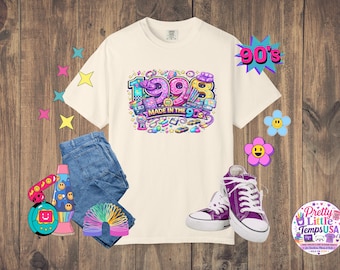 Könnte beinhalten: Ein cremefarbenes Kurzarm-T-Shirt mit einer bunten Grafik und dem Text 1998 MADE IN THE 90S, umgeben von einer blauen Jeans, violetten Stoffturnschuhen, einer Regenbogen-Spirale, einer Lavalampe, einem digitalen Haustier sowie Stern- und Blumenstickern.