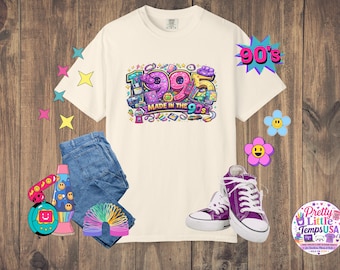 Könnte beinhalten: Ein cremefarbenes Kurzarm-T-Shirt mit einer bunten Grafik und dem Text 1995 MADE IN THE 90S, umgeben von Illustrationen eines Kaugummiautomaten, eines Handheld-Spiels, einer Lavalampe, einer Regenbogenspirale sowie lila Turnschuhen und blauen Jeans.