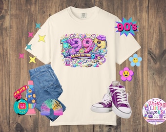 Könnte beinhalten: Ein cremefarbenes T-Shirt mit einer bunten Grafik der Jahreszahl 1999 und dem Text MADE IN THE 90S, umgeben von Illustrationen von Retro-Spielzeug, einer blauen Jeans, violetten Stoffturnschuhen und verschiedenen dekorativen Aufklebern wie Blumen und Sternen.