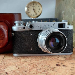 Puede incluir: Una cámara telemétrica FED-2 plateada y negra con objetivo, situada junto a un estuche de cuero marrón con el texto FED-2 grabado. Un pequeño reloj analógico redondo con esfera blanca y manecillas negras se sitúa tras la cámara.