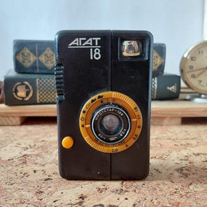 May include: A black Agat 18 film camera featuring a prominent orange circular lens dial with aperture and weather symbols. The camera body displays the text Agat 18 and Industar-104, with a small orange shutter button on the front panel.