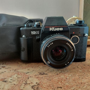 Op de afbeelding: Een zwarte Kiev 19M 35mm filmcamera met een 50mm Arsat H lens met een 1:2 diafragma. De camerabehuizing heeft een gestructureerde grip en staat naast een zwarte cameratas. De merknaam Kiev staat in witte letters op de voorzijde van de camera.