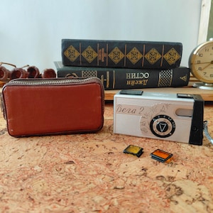 May include: A silver Vega 2 film camera with a black side panel, a brown leather zippered case, and two small square colored filters. The camera features engraved line art and text on its front face, positioned near two stacked dark books.