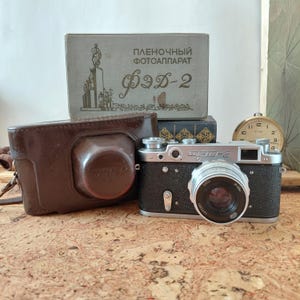 May include: A silver and black FED-2 rangefinder camera with a lens, positioned next to its brown leather case. Behind the camera sits a gray cardboard box with the text "ПЛЕНОЧНЫЙ ФОТОАППАРАТ ФЭД-2" and a graphic illustration of a monument.