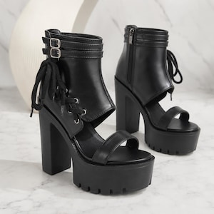 May include: A pair of black platform ankle boots featuring a high block heel, a thick lug sole, and an open-toe design. The boots include side cutouts, black lace-up detailing, and two adjustable buckle straps around the ankle for a secure fit.