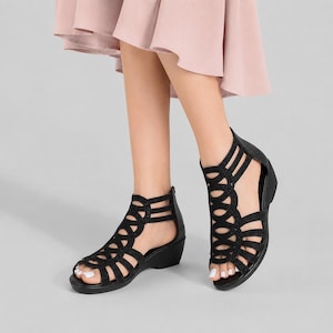 May include: Black sandals featuring a cage-style upper with circular cutouts and a low wedge heel. The footwear has a back zipper closure and a textured finish on the straps, providing a secure fit for the foot while maintaining an open design.