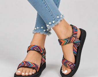 May include: A pair of sandals featuring black soles and adjustable straps with a vibrant, multi-colored geometric pattern in shades of pink, blue, yellow, and orange. The straps secure around the ankle and across the forefoot with hook-and-loop closures.