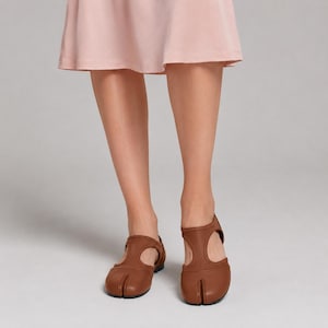 May include: A pair of brown leather split-toe shoes featuring a T-strap design with a rounded heel and a smooth finish, worn with a light pink skirt that falls just above the knees, showcasing a distinct separation between the big toe and other toes.