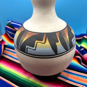 May include: A matte off-white ceramic vase with a rounded body and a narrow neck. The midsection features a horizontal band decorated with black geometric shapes over a gradient of orange, yellow, and blue colors. The rim is painted black.