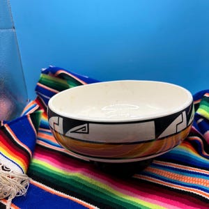 May include: A round ceramic bowl with a cream-colored interior and a black rim featuring white geometric patterns. The exterior base displays a warm, marbled gradient of orange, brown, and yellow tones, creating a distinct contrast.