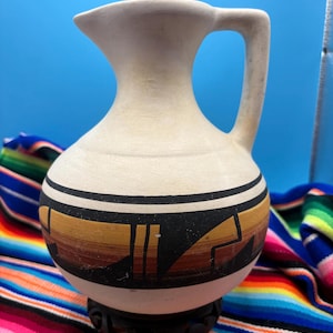 May include: A cream-colored ceramic pitcher with a single handle and a wide, rounded body. The vessel features a horizontal band decorated with black, brown, and tan geometric patterns encircling the midsection, resting on a dark base.
