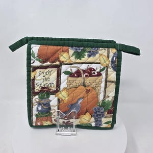 May include: A square quilted potholder with a dark green border and hanging tabs. The fabric features a harvest-themed print with pumpkins, a basket of cherries, grapes, and a blue bird. The text "Enjoy the Season" is printed on a cream square.