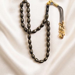 Peut inclure: Un chapelet composé de perles noires de forme ovale avec des rayures verticales dorées. Le cordon se termine par un ornement décoratif en métal doré et un pompon à plusieurs brins argentés agrémenté de petites breloques circulaires.