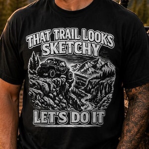 Könnte beinhalten: Ein schwarzes Kurzarm-T-Shirt mit einem weißen Grafikdruck eines Geländewagens auf einem felsigen Bergpfad. Der Text lautet That Trail Looks Sketchy Let's Do It. Das Design enthält detaillierte Illustrationen von Kiefern und Bergen.