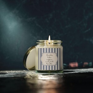 May include: A clear glass candle jar with a lit wick and a gold-toned metal lid resting beside it. The label features a blue and white vertical striped pattern with a central white frame containing the text Smells like coffee and plot twists.