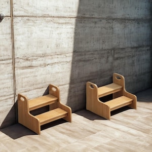 Puede incluir: Dos taburetes de madera de tono claro, cada uno con dos escalones horizontales y paneles laterales con aberturas ovaladas para las manos. Los taburetes están fabricados en madera natural lisa con veta visible, sobre una superficie plana.
