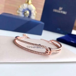 Puede incluir: Una pulsera en tono oro rosa con un diseño de doble banda y un detalle de nudo central. Las bandas están incrustadas con pequeños cristales transparentes. La pulsera incluye una delicada cadena de extensión y el logo Swarovski.