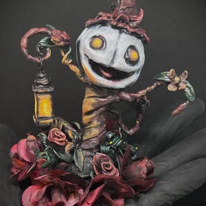 Könnte beinhalten: Eine skulptierte Figur mit einem blassen, runden Gesicht, leuchtend gelben Augen und einem breiten Grinsen, die einen dunkelroten Hut trägt. Die Figur ist von dunkelroten Rosen umgeben und hält eine kleine, gelb leuchtende Laterne.