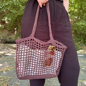 May include: A dark mauve crochet tote bag featuring an open grid pattern and two shoulder straps. The bag contains a white fabric liner and has a pair of amber-tinted square sunglasses hanging from the front of the crocheted mesh exterior.