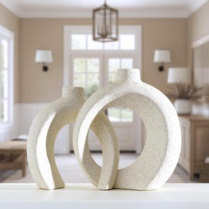 May include: Two speckled, off-white ceramic vases with a modern, open-circle design. The vases have a textured surface and a neutral color palette, suitable for home decor. The vases are displayed on a white surface.
