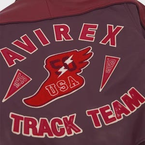 以下が含まれることがあります： 赤と白の刺繍で「AVIREX TRACK TEAM」と大きく記された、ダークマルーンカラーのレザージャケット。中央の翼のエンブレムには数字の50、USAの文字、そして1975と2025と記された2つの三角形のペナントがデザインされています。