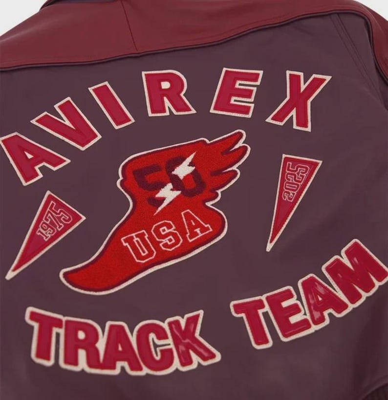 以下が含まれることがあります： 赤と白の刺繍で「AVIREX TRACK TEAM」と大きく記された、ダークマルーンカラーのレザージャケット。中央の翼のエンブレムには数字の50、USAの文字、そして1975と2025と記された2つの三角形のペナントがデザインされています。